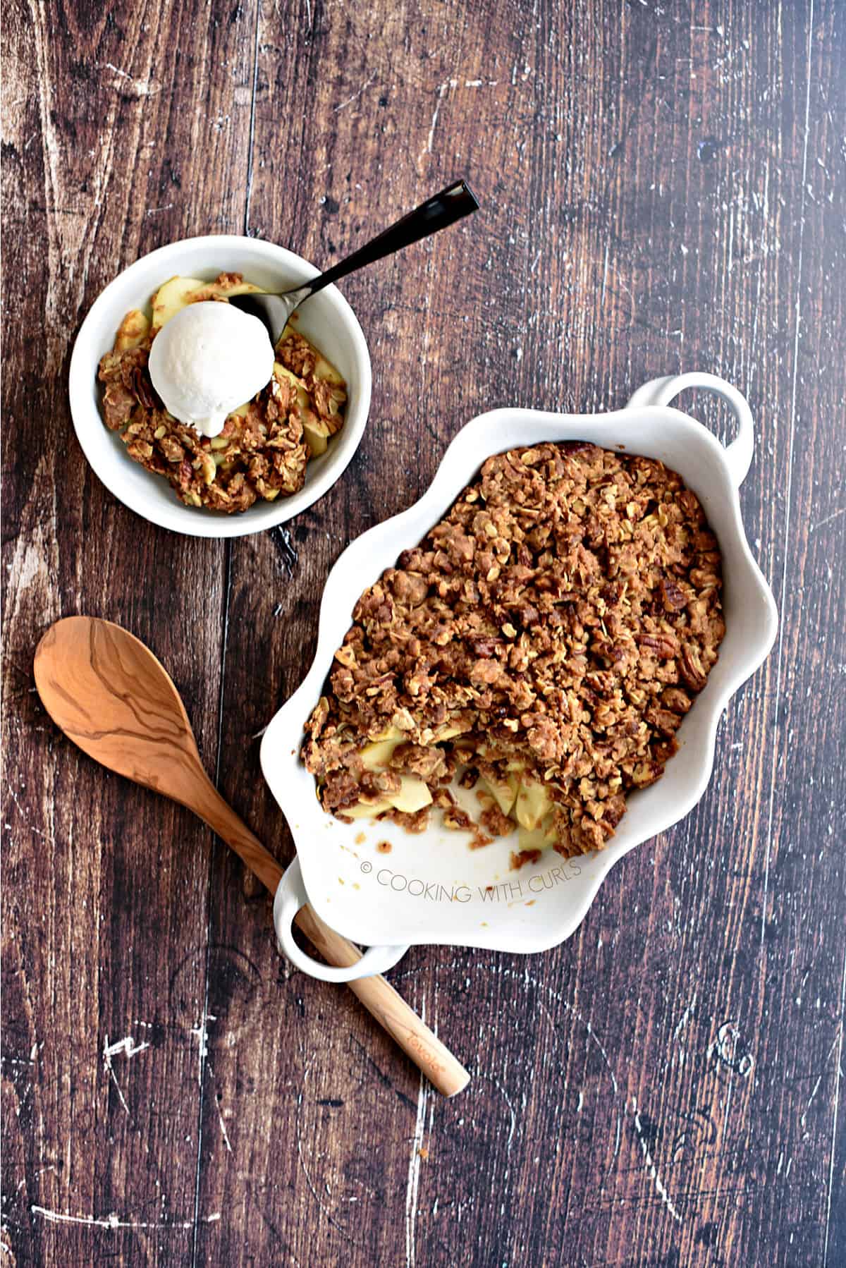 looking down on a baked apple crisp in the baking dish, a wooden spoon laying next to it and a white bowl with a scoop of ice cream on top of the apple crisp.