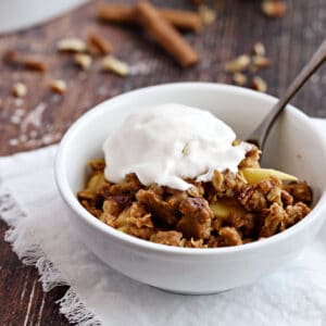 A white bowl with apple crisp topped with vanilla ice cream sitting on a white fringed napkin with an apple and baking dish in the background.