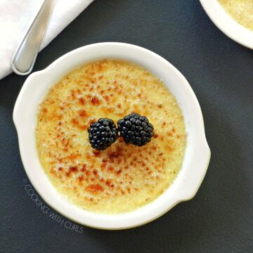 Two white dishes with creme brulee topped with blackberries, with a white napkin and two spoons in the upper left corner.