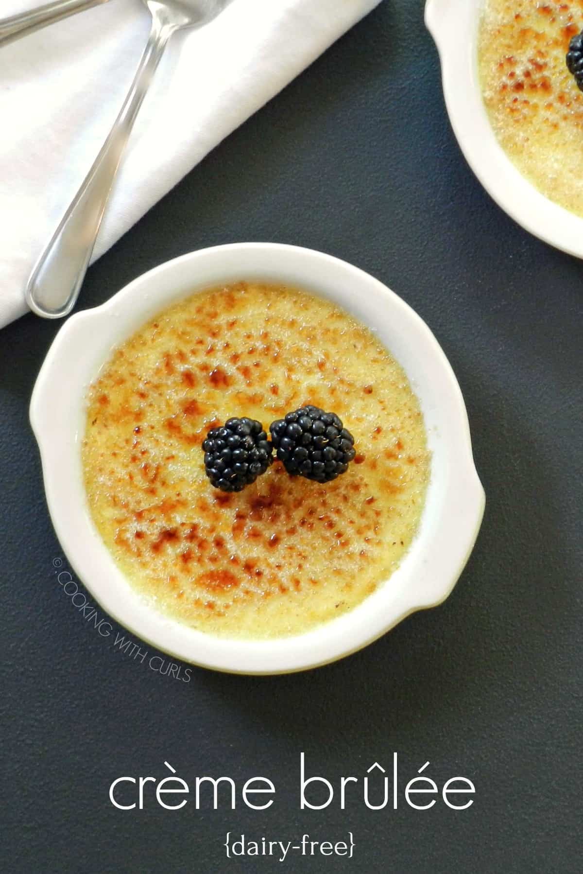 Two white dishes with creme brulee topped with blackberries, with a white napkin and two spoons in the upper left corner.
