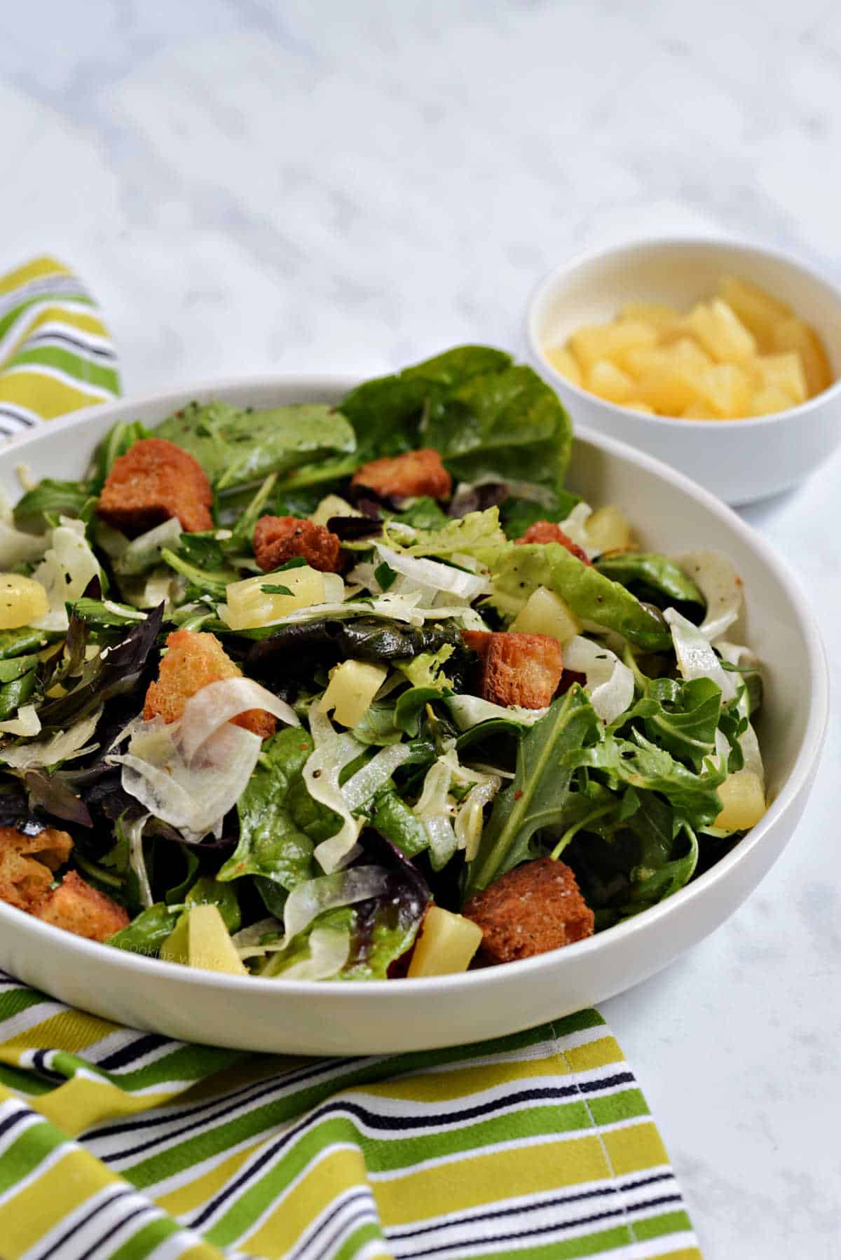 Shaved fennel salad with mixed greens, pineapple chunks and croutons with a small bowl of pineapple in the background.