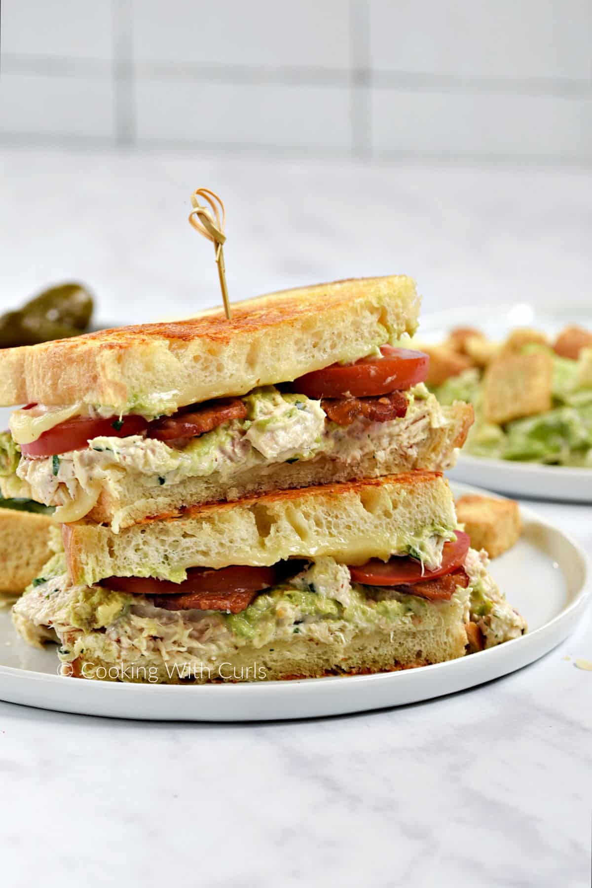 Two halves of a tuna melt sandwich with bacon, avocado, and tomatoes stacked on a plate with a Caesar salad. 