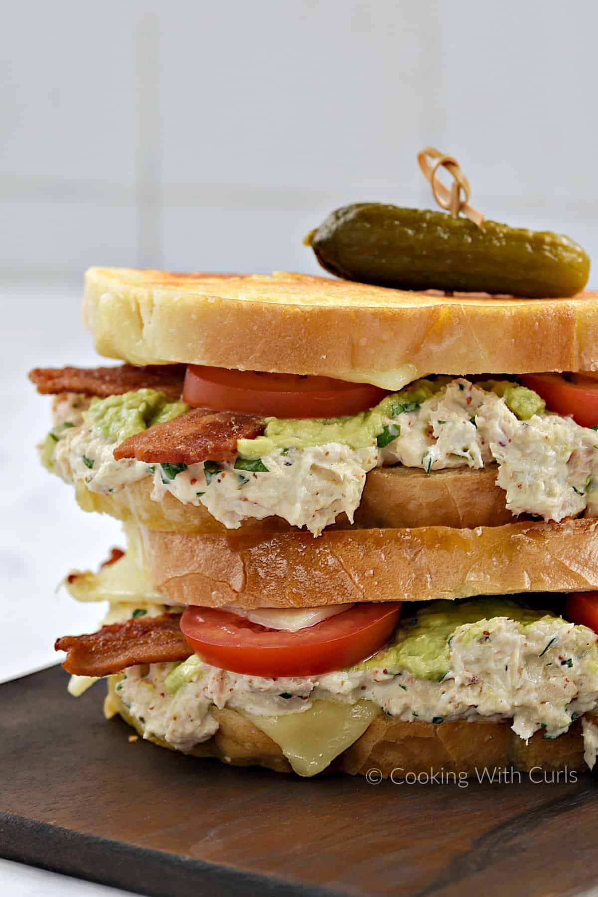Two tuna melt sandwiches with bacon, tomato, cheese, and avocado, stacked on top of each other with a pickle on top.
