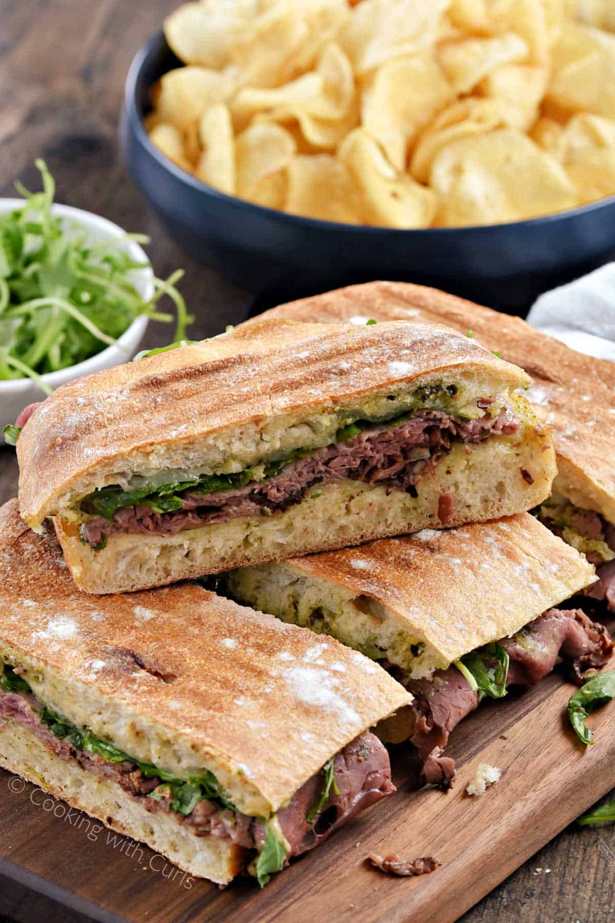 Roast beef panini sandwiches stacked on top of each other on a wooden board with potato chips in the background.
