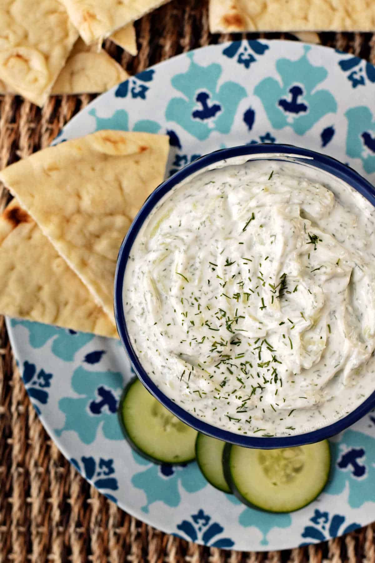 Greek Tzatziki dip in a small bowl surrounded by pita chips and sliced cucumbers.
