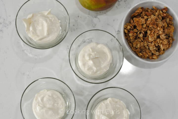Greek yogurt in four small glass bowls with a mango and bowl of granola on the side.