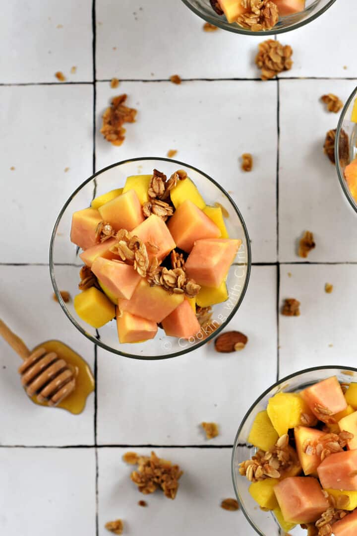 Looking down on papaya and mango chunks sprinkled with granola in four small dessert glasses.