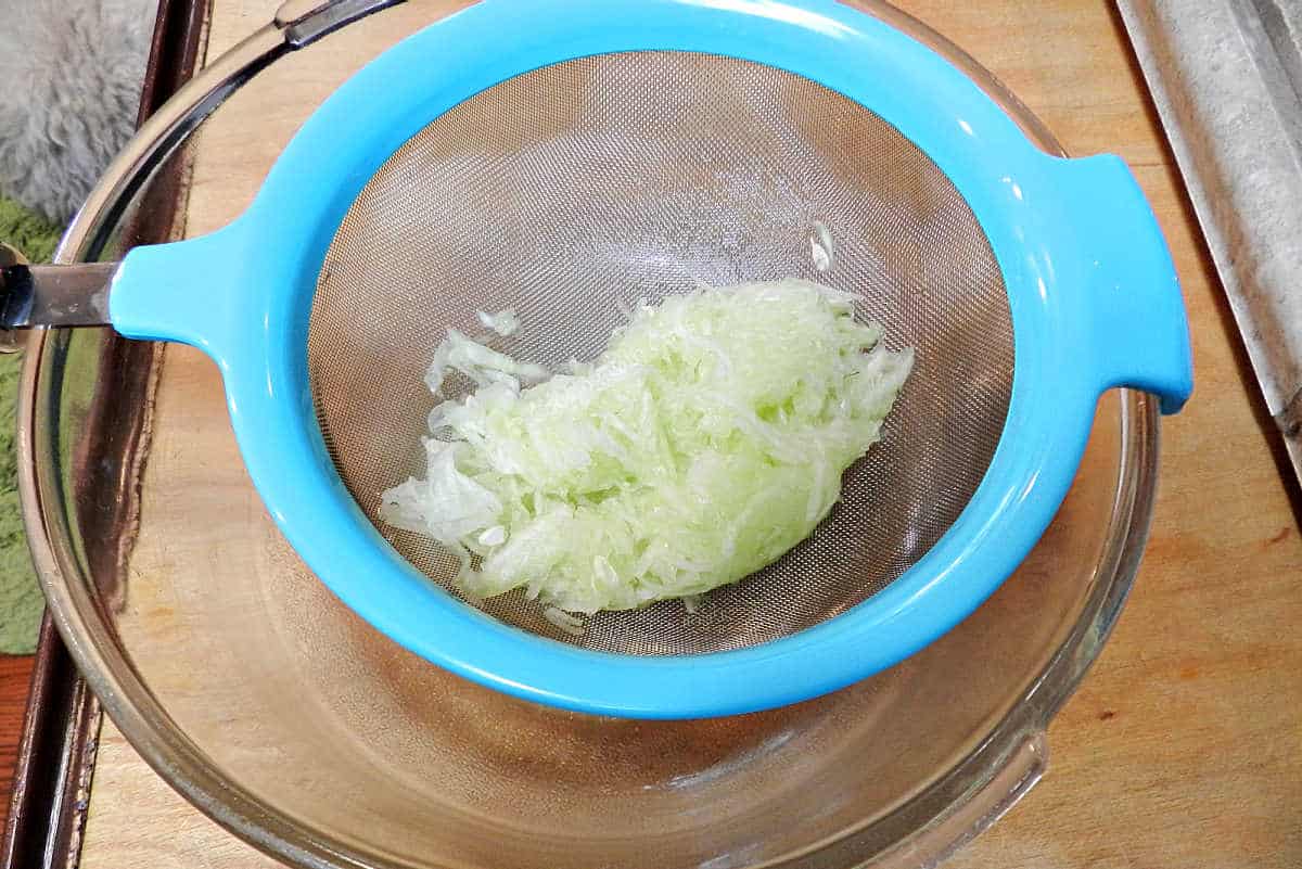Shredded cucumber in a strainer over a large bowl.