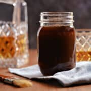 Whiskey glaze in a glass jar with a bottle and glass of whiskey in the background.