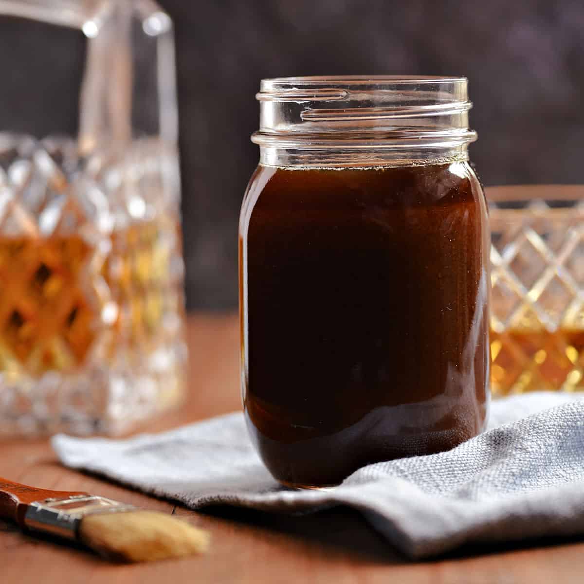 Whiskey glaze in a glass jar with a bottle and glass of whiskey in the background.