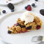 a slice of apple-berry crumble topped with irish whiskey whipped cream sitting on a white plate with a fork laying on the edge and mixed berries laying behind.