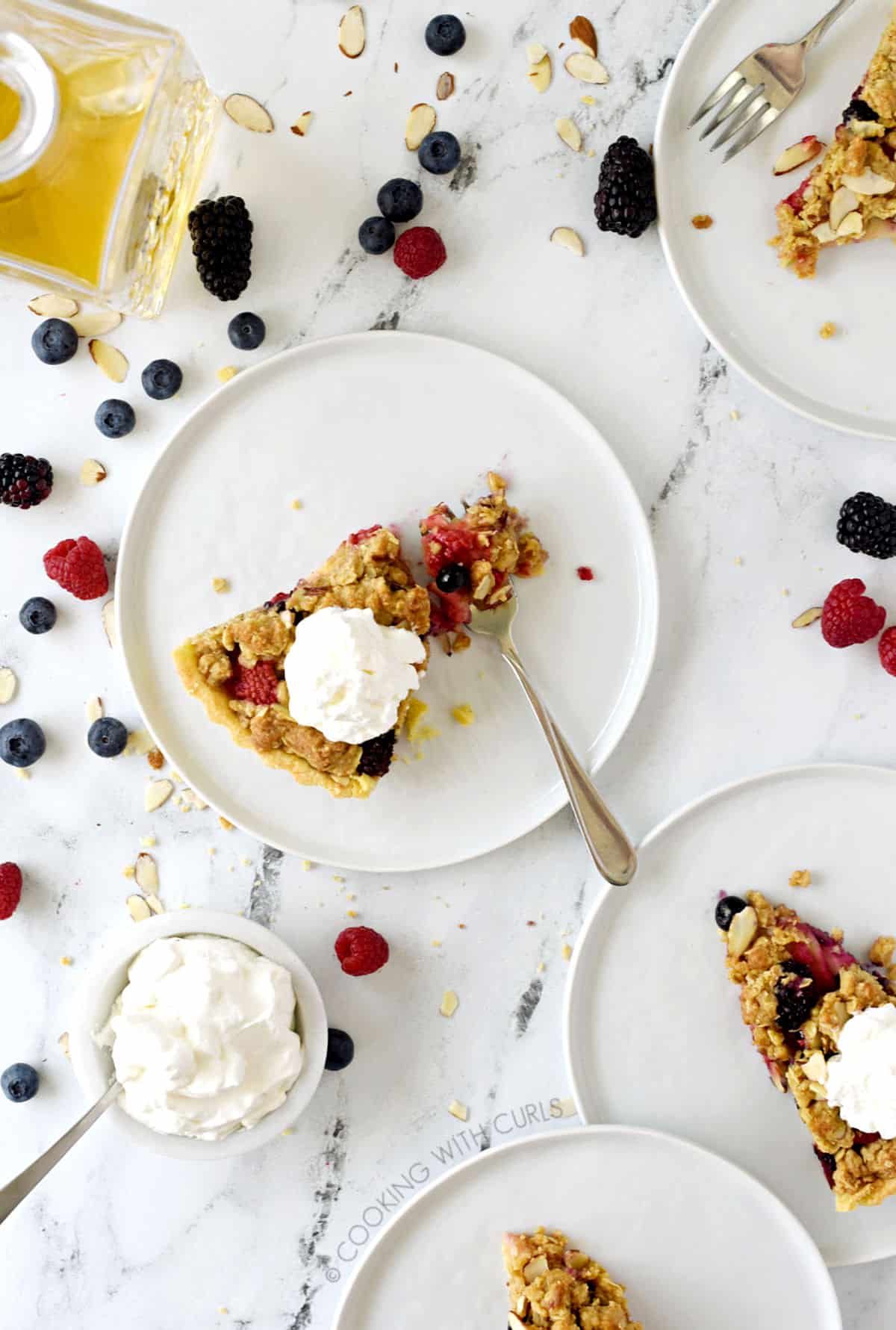 looking down on four, small white plate topped with apple-berry crumble and topped with whipped cream with mixed berries scattered around the marble background and a small white bowl of whipped cream in the lower left corner.