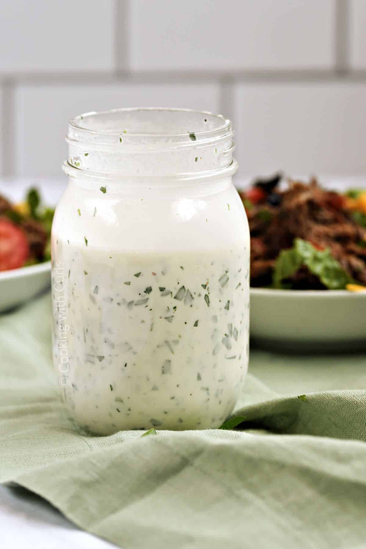 Dairy Free Ranch Dressing in a Mason Jar sitting on a green napkin with two salads in the background. 