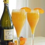 Two champagne glasses filled with Peach Bellini with a bottle of prosecco in the background.