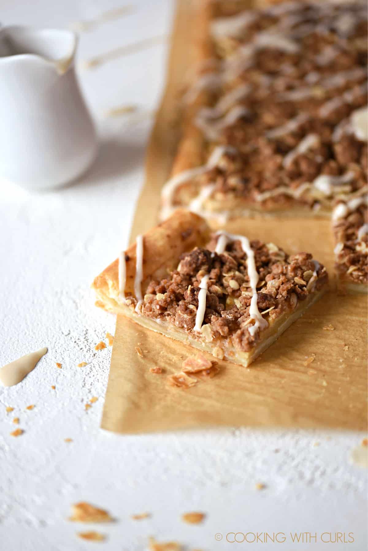 an apple streusel pizza on a sheet of parchment paper with a piece pulled out at an angle with a pitcher of glaze in the background.