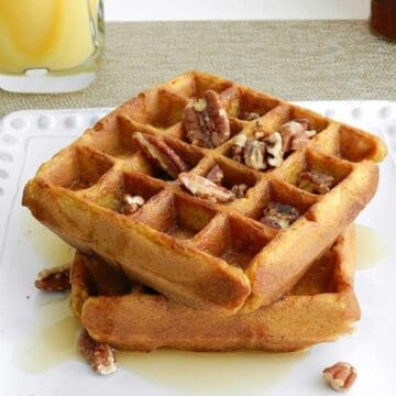 Two pumpkin Belgian waffles topped with maple syrup and pecans stacked on a plate.