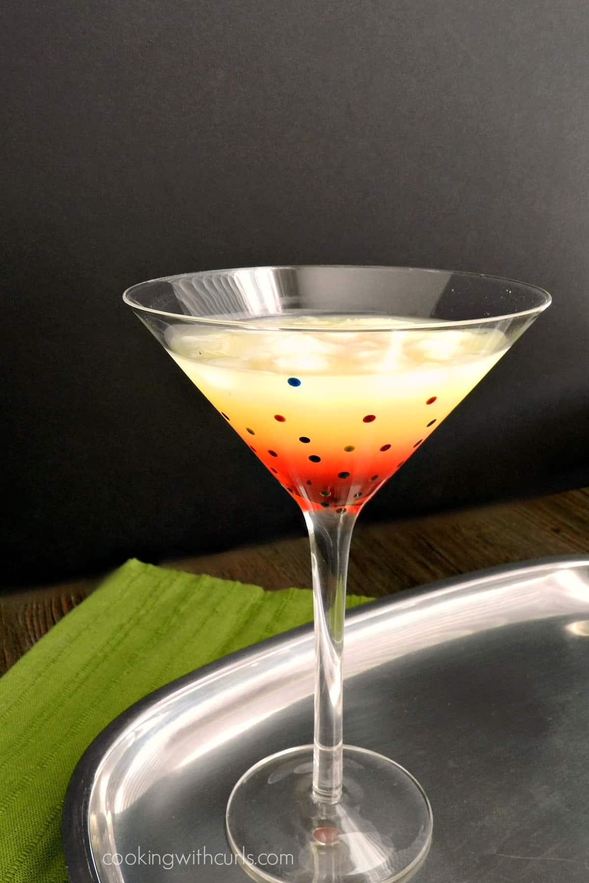 A tequila sunrise cocktail in a martini glass sitting on a silver tray with a green napkin behind it.