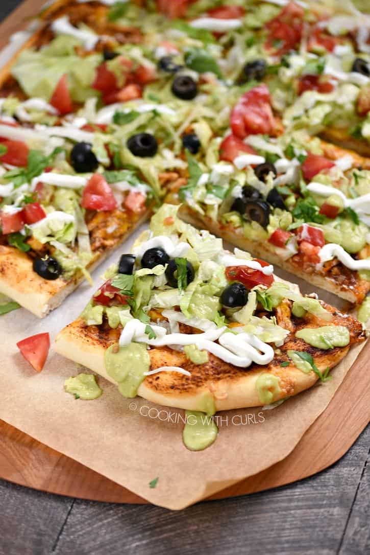 A rectangle pizza topped with lettuce, chicken, tomatoes, olives, guacamole, and sour cream cut into squares.