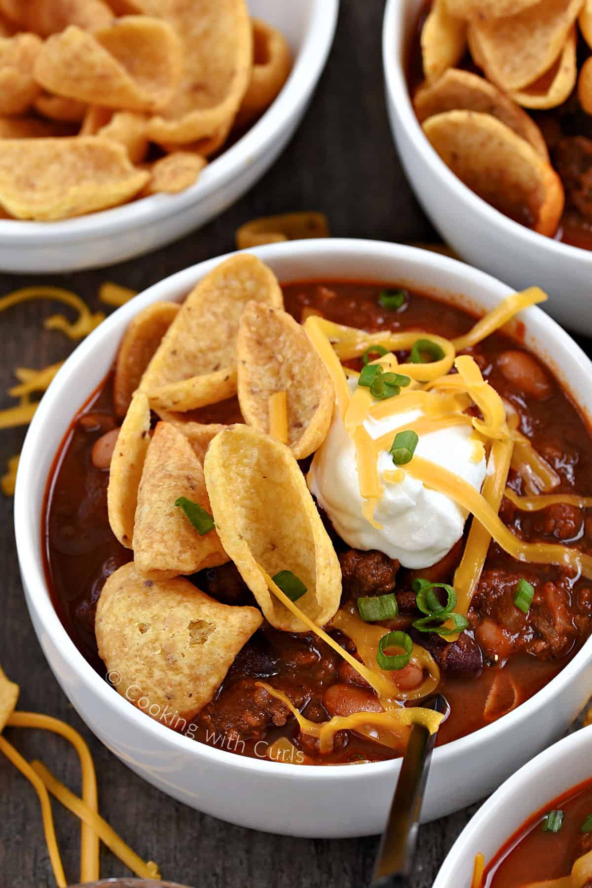 A bowl of Jack Daniels Chili topped with sour cream, cheddar cheese, green onions, and corn chips with additional bowls in the background. 