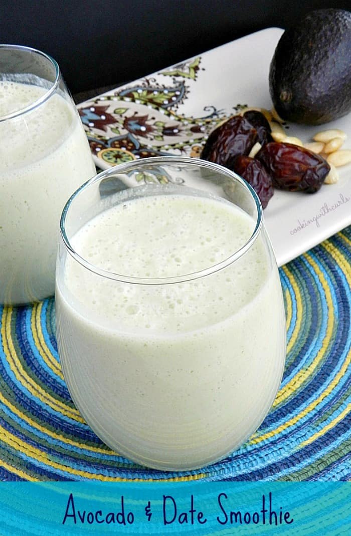 Avocado and Date Smoothie in a small glass sitting on a blue and green spiral placemat, with a plate of dates, avocado and almonds in the background