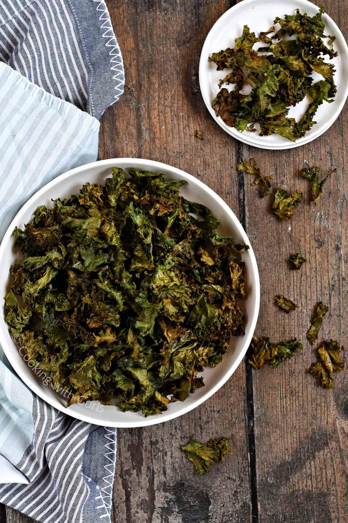 Looking down on a bowl of crispy kale chips with scattered chips on the table leading to a small plate.