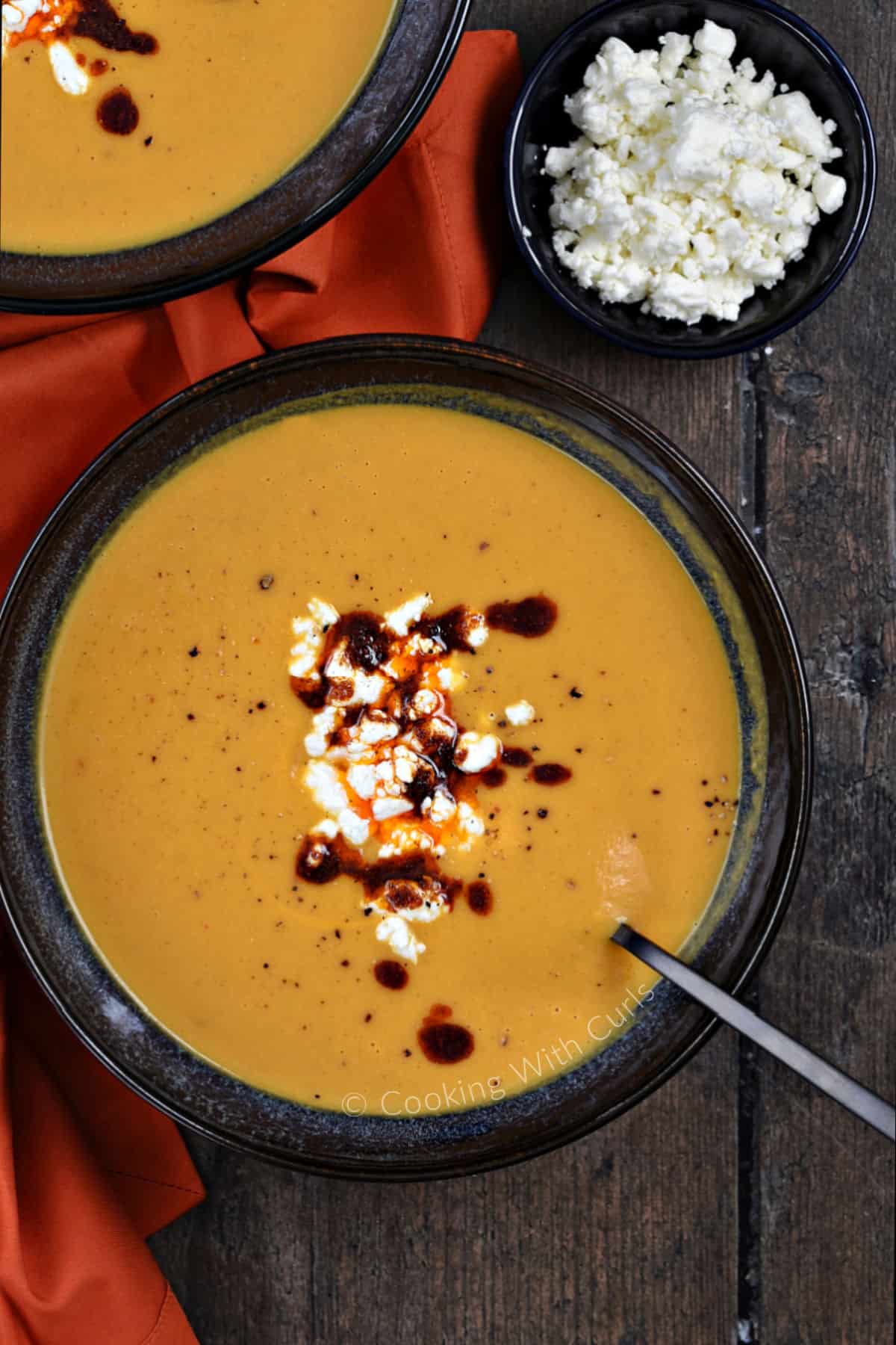 Looking down on two bowls of bright orange soup with crumbled feta and chili sauce in the center and a small bowl of feta cheese on the side. 