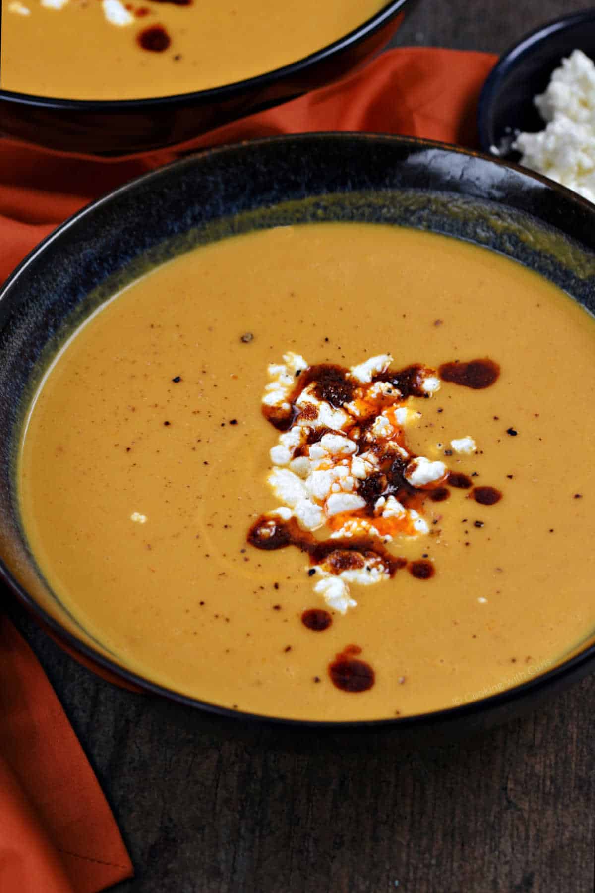 Two bowls of orange soup with feta cheese and chili sauce in the center.