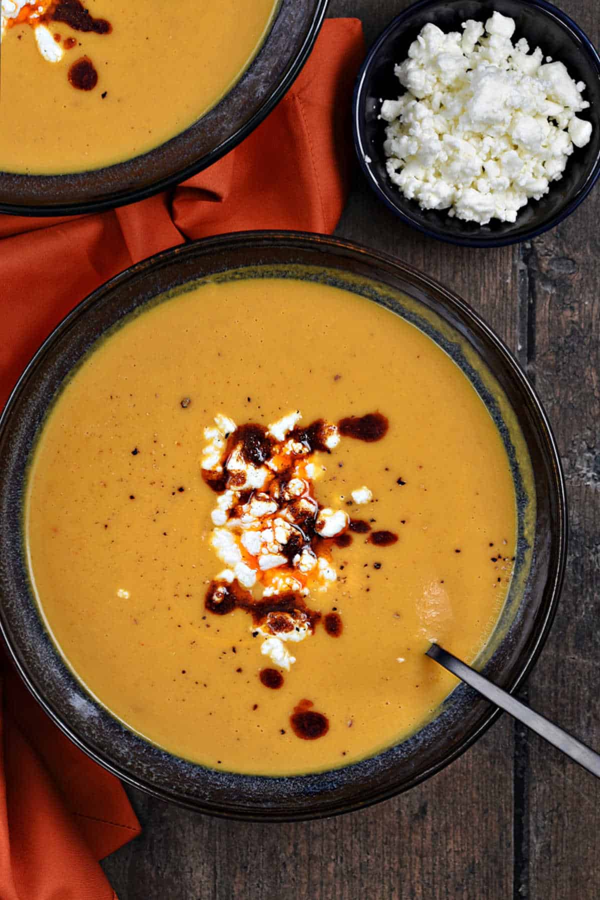 Two bowls of orange soup with feta cheese and chili sauce in the center.