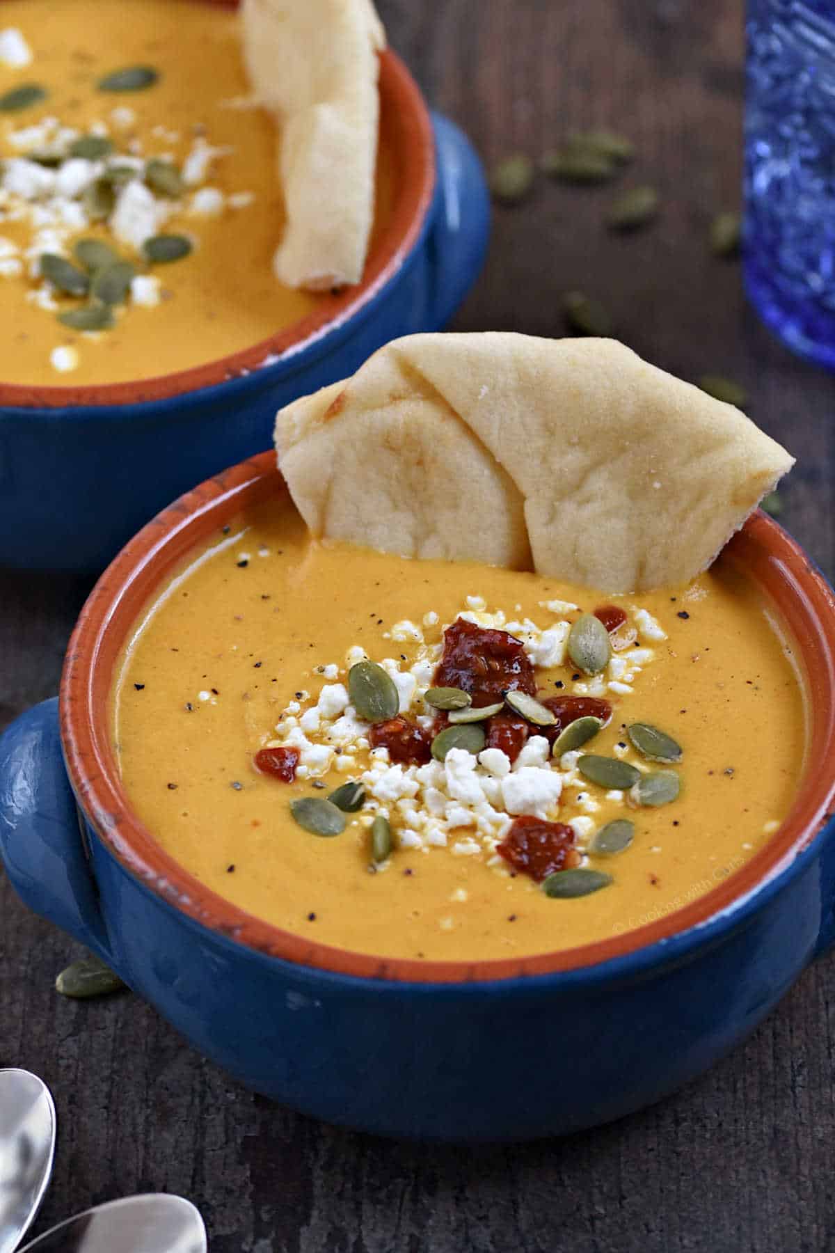 Two blue bowls of orange soup with pita bread, feta cheese, and pumpkin seeds.