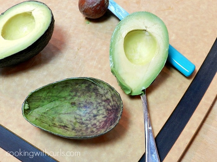 avocado flesh scooped out with a spoon resting on a cutting board with the avocado pit, blue knife and other half in the background 