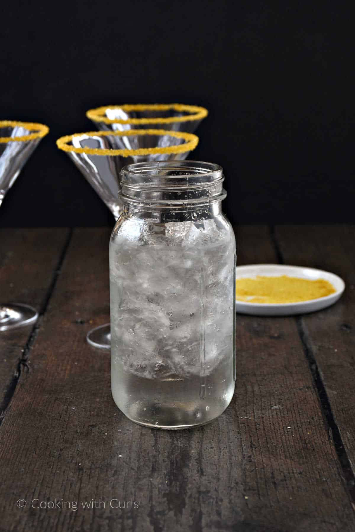 A glass cocktail shaker filled with ice, caramel vodka, apple vodka, and butterscotch schnapps with three martini glasses in the background.