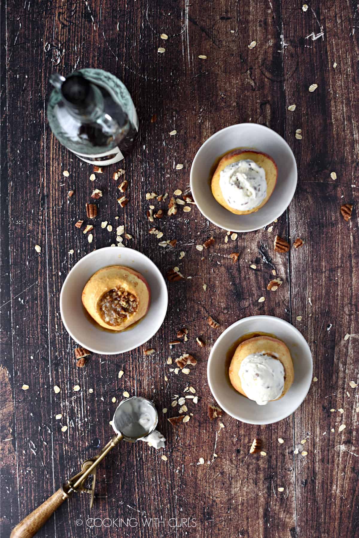 looking down on three small white bowls with spiced rum baked apples, two topped with ice cream. there is an ice cream scoop in the lower left corner and a bottle of spiced rum in the upper left corner.