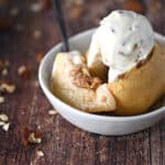 a stuffed, baked apple with a slice cut out and topped with ice cream sitting in a small white bowl with a black spoon sticking out the left side, surrounded by oats and pecans.