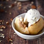 a stuffed, baked apple with a slice cut out and topped with ice cream sitting in a small white bowl surrounded by oats and pecans.
