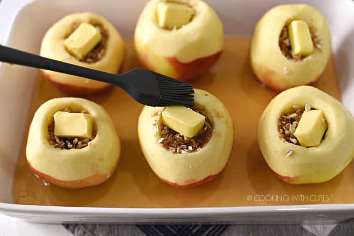 six stuffed apples topped with butter in a white baking dish with spiced rum and apple cider being basted with a gray silicone brush. 