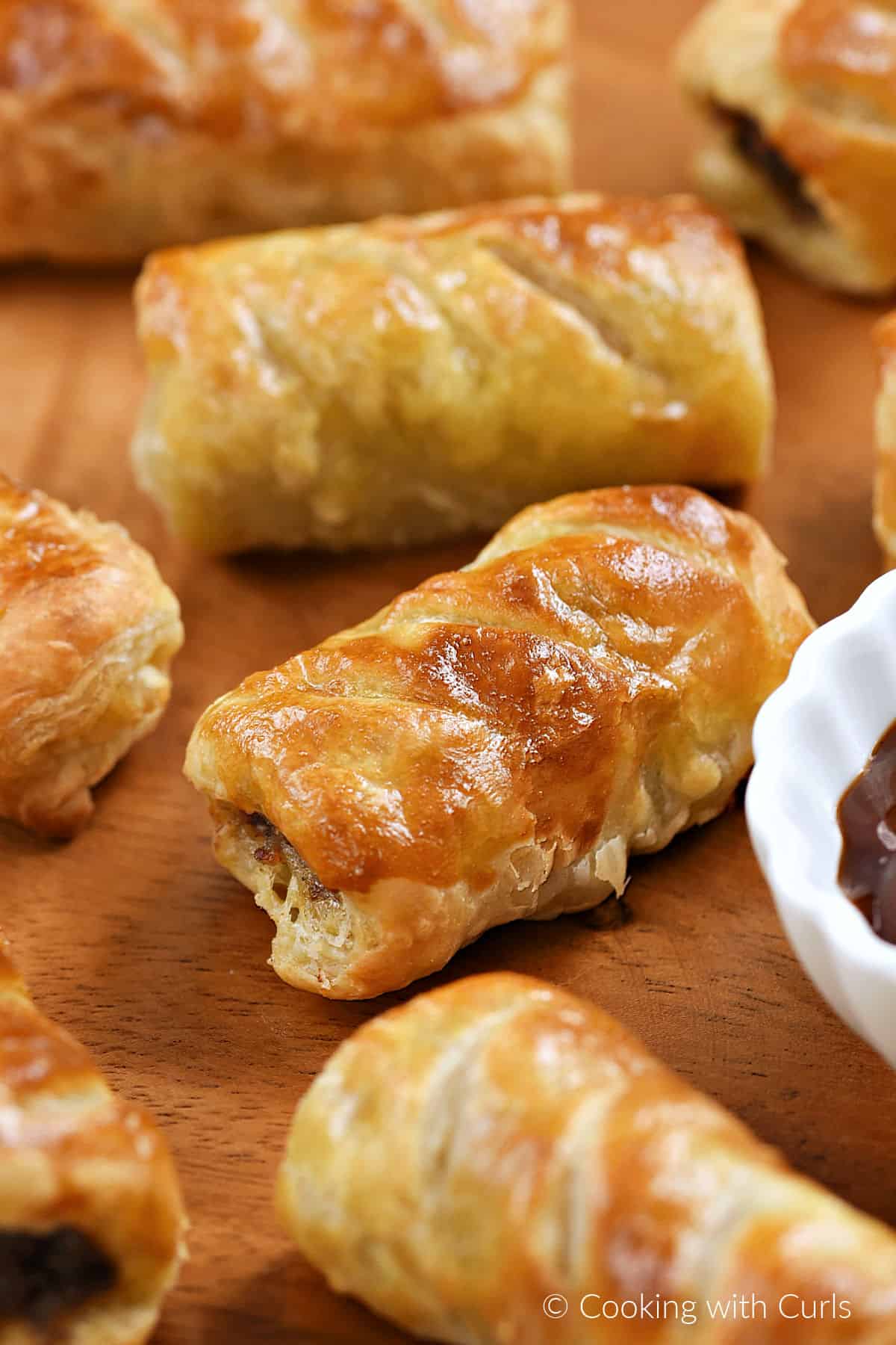 Eight sausage rolls on a wooden tray with a bowl of sauce. 