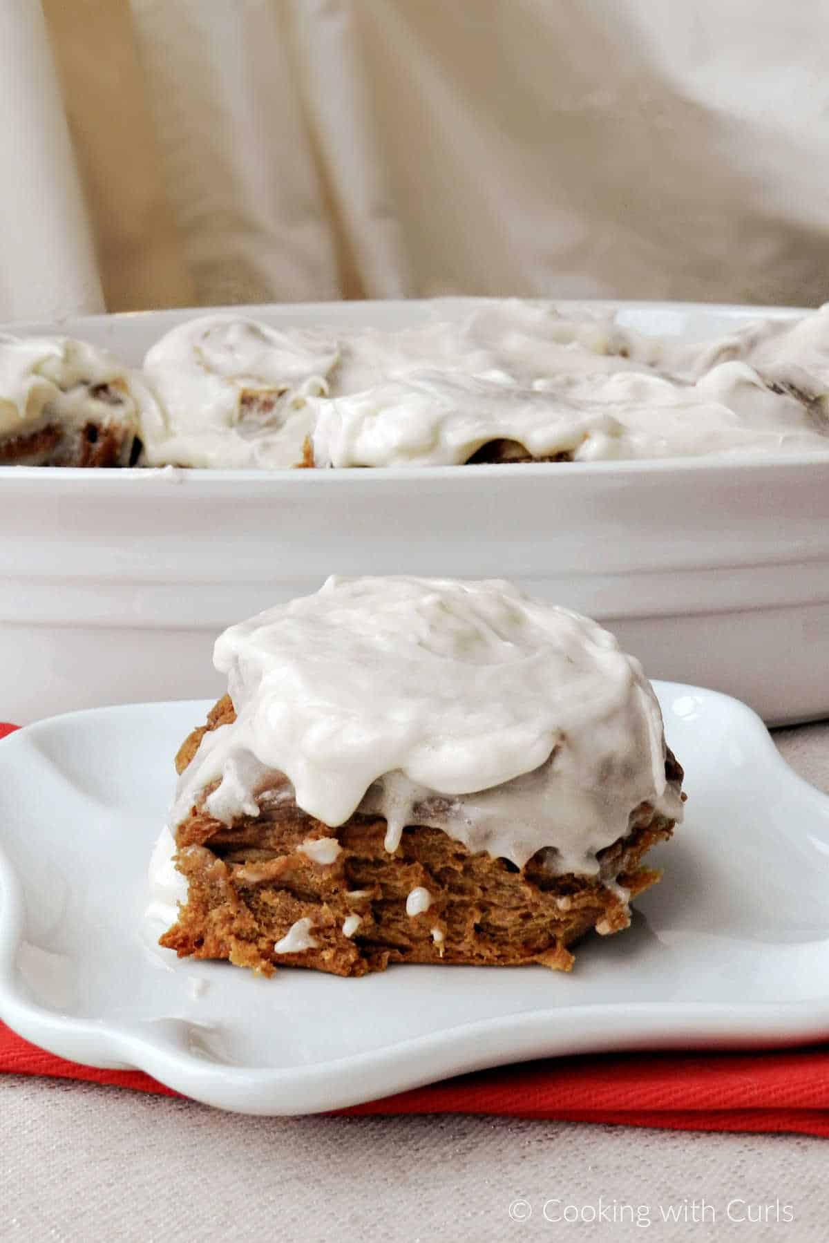 A gingerbread cinnamon rolls sitting on a plate with a pan of rolls in the background.