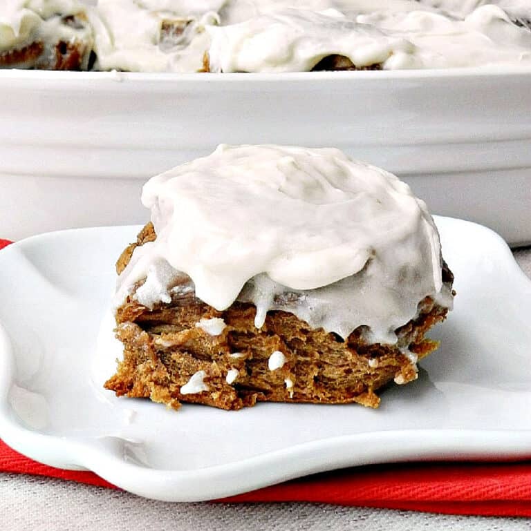 Gingerbread cinnamon roll on a white plate sitting on a red napkin with a pan of rolls in the background.