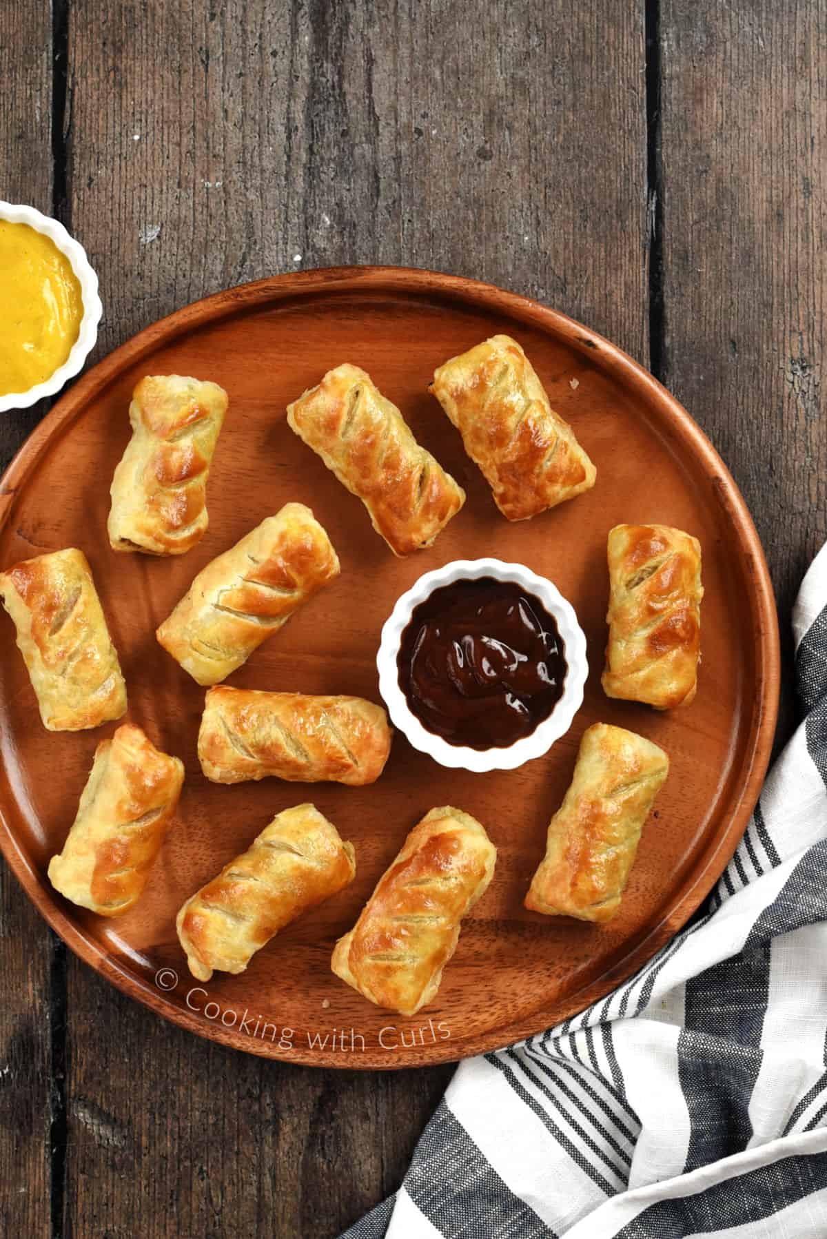 Looking down on eleven sausage rolls on a wood platter with a bowl of sauce and a bowl of mustard. 