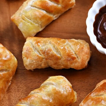 Looking down on five sausage rolls on a wood platter with a small bowl of sauce.