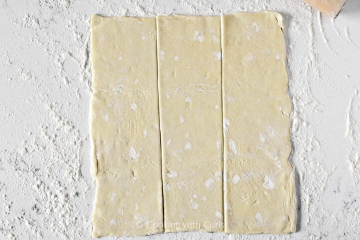 Puff pastry dough rectangle cut into three strips on a floured work surface. 