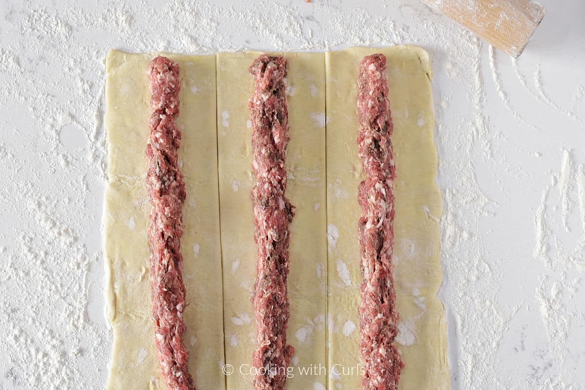 Three strips of puff pastry dough with sausage running down the center on a floured surface. 