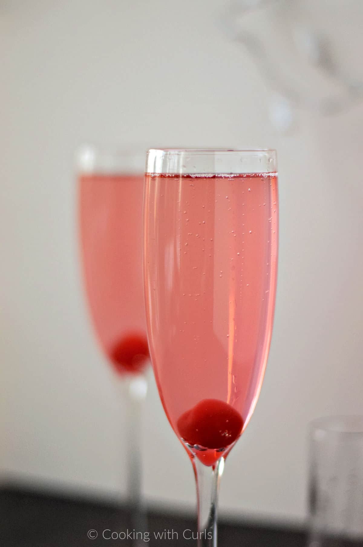 Two pink, sparkling cocktails with a cherry at the bottom of a champagne flute.