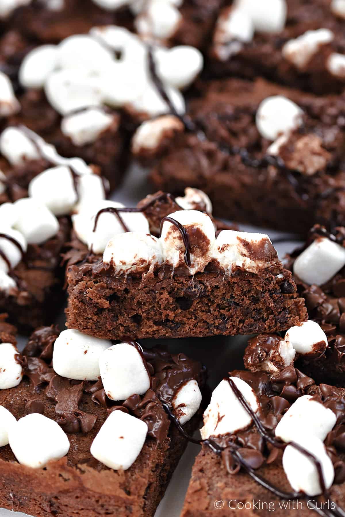 A single chocolate brownie topped with melted chocolate and mini marshmallows resting on top of a platter full of brownies. 