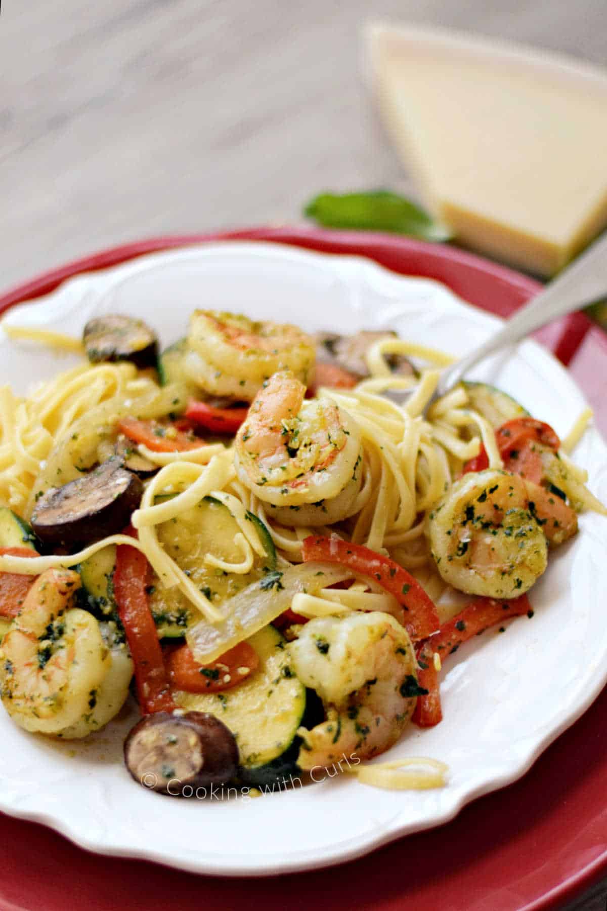 Pesto shrimp, bell pepper, zucchini, mushrooms and pasta on a plate with a wedge of parmesan in the background.