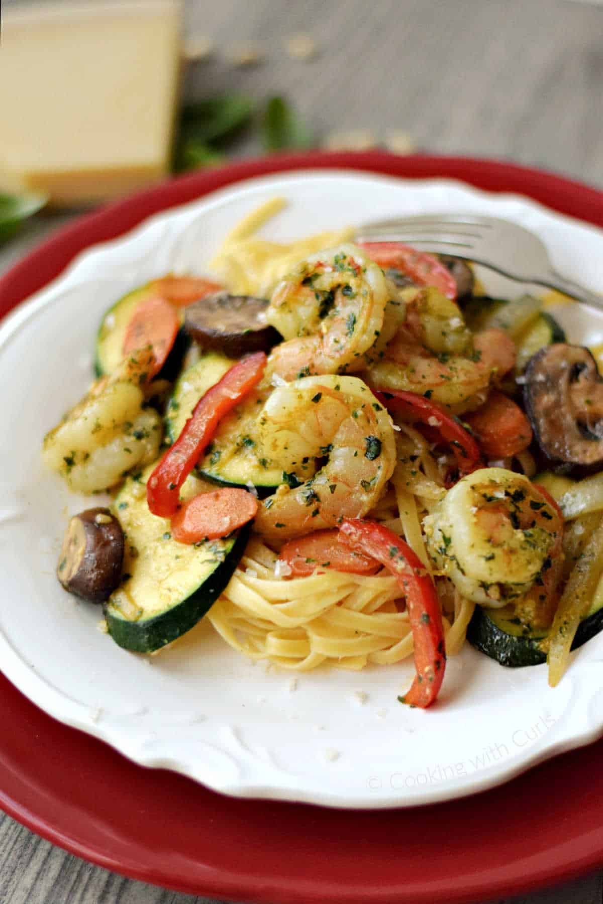 Pesto shrimp, bell pepper, zucchini, mushrooms and pasta on a plate with a wedge of parmesan in the background.