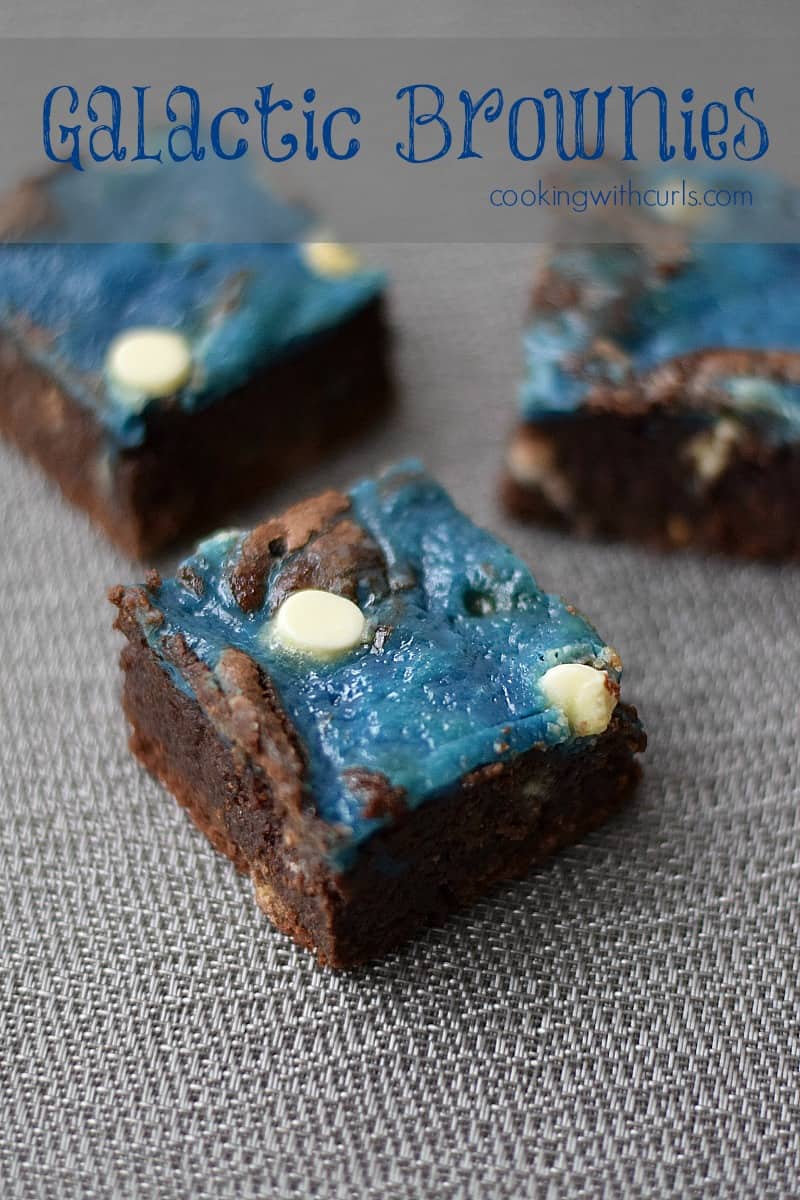 Three blue swirled cream cheese brownies topped with white chocolate chips on a silver metalic mat.