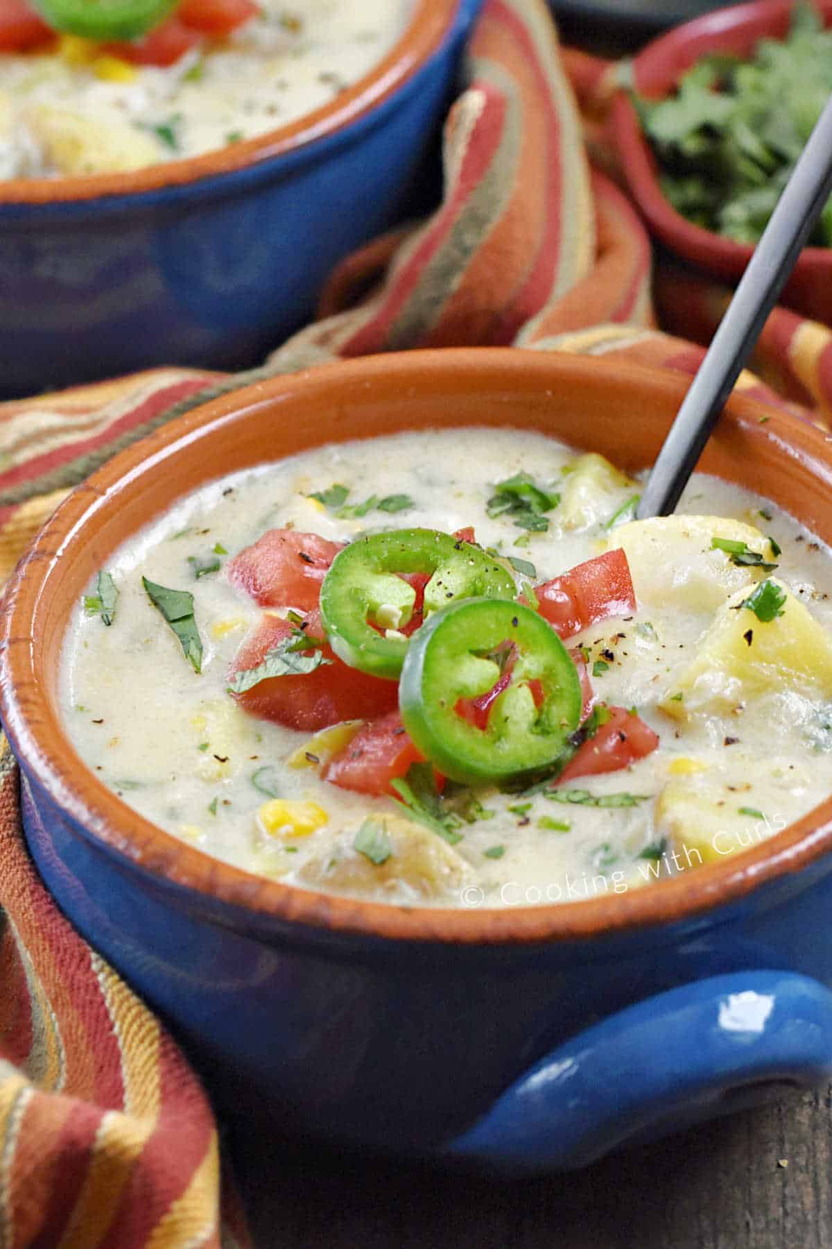 A bowl of Southwest Crab Chowder topped with sliced jalapenos, diced tomato, and chopped cilantro with a black spoon in the upper right corner. 