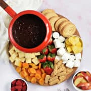 Chocolate fondue in pot surrounded by fruits, cookies, and marshmallows.