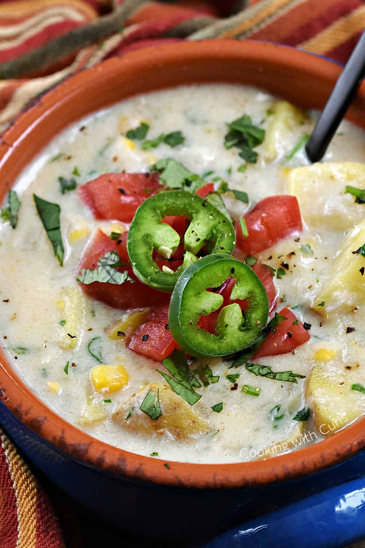 Close-up of sliced jalapenos, diced tomatoes, and cilantro on top of a bowl of crab chowder with potatoes and corn. 